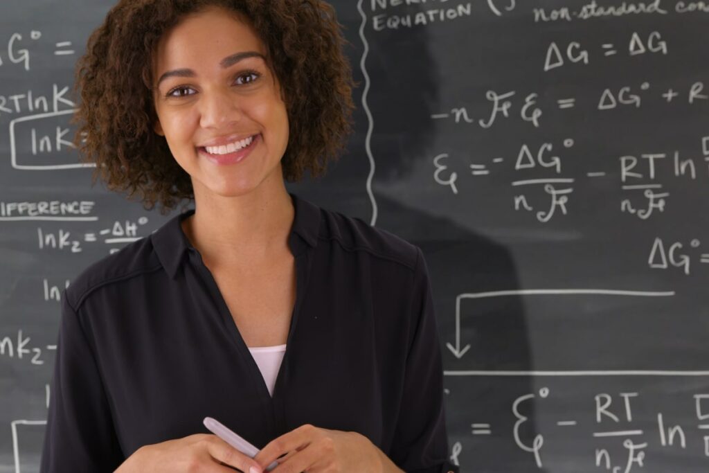 Math teacher with a chalk board full of math equations in the background. She's smiling at the camera and wearing a black shirt.