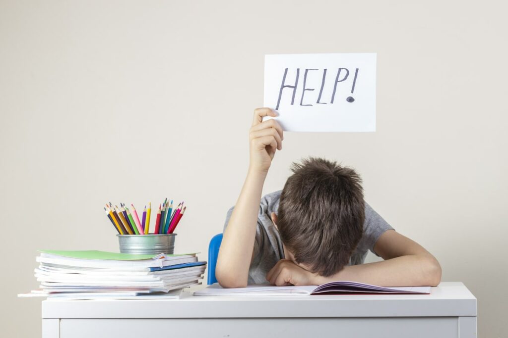 Child with his head down on desk. Books and pencils next to him. He's holding up a sign that says "help"